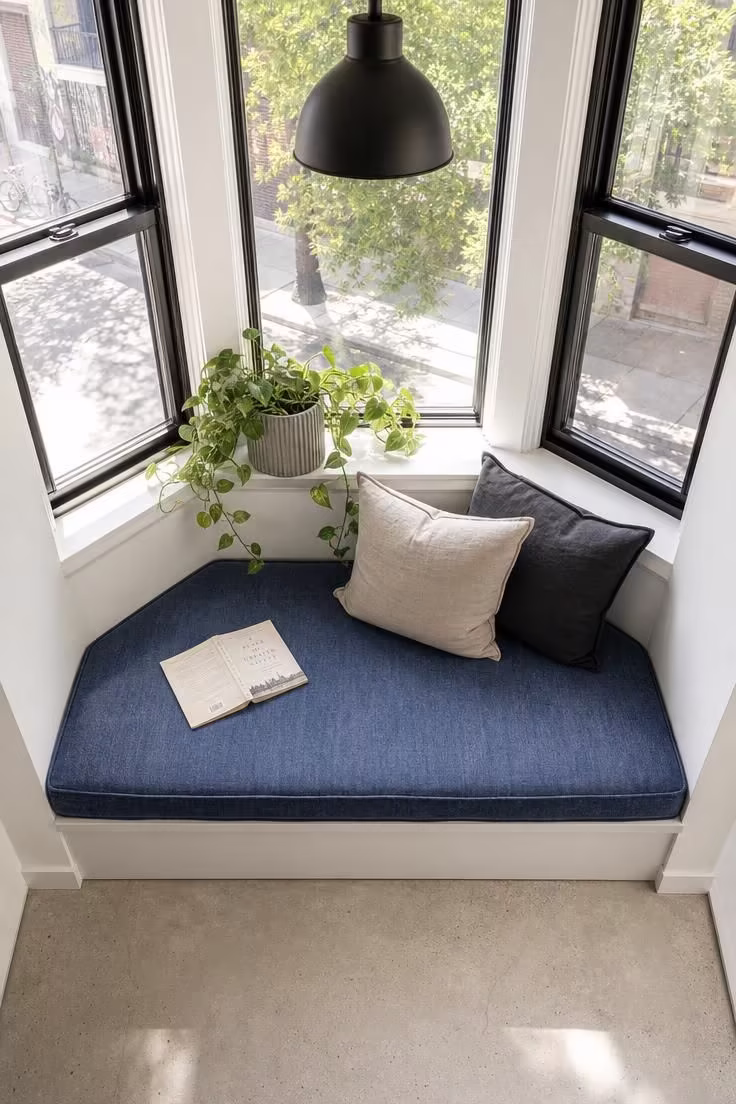 Overhead view of a bay window reading nook with denim cushion, ribbed concrete planter, and matte black pendant in bright midday light.