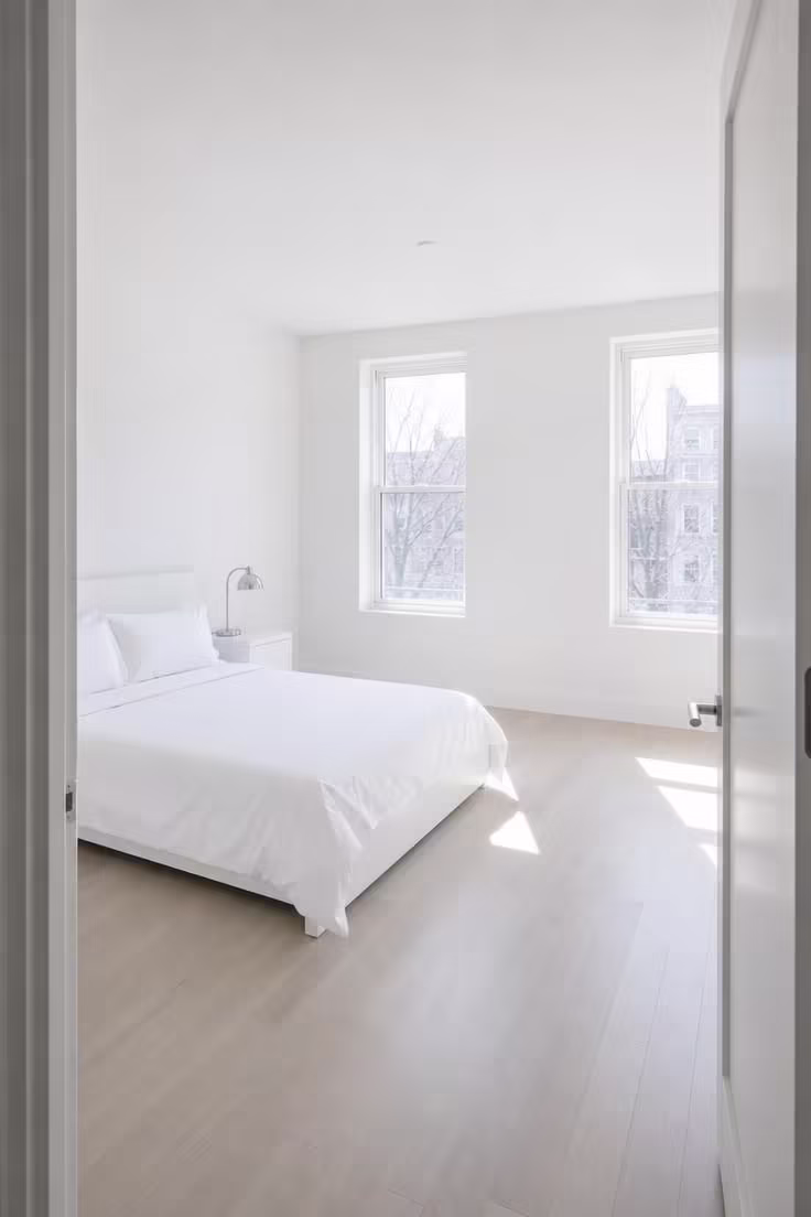 A stark white bedroom under bright midday light showing how flat white walls and minimal layering create an empty, clinical feeling.