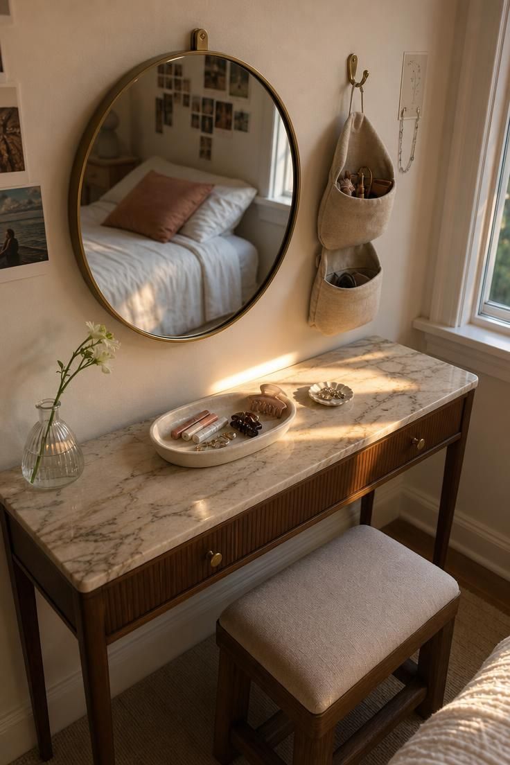 Small round brass-framed vanity mirror above a marble-top table with linen pouch organizers and a wooden stool