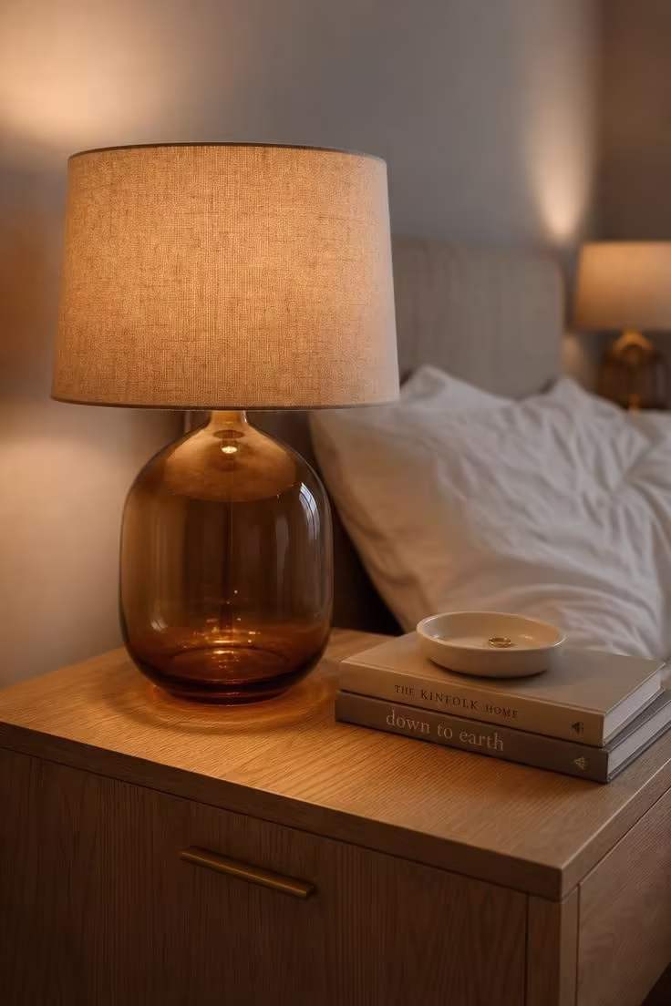 13 Cozy Bedroom Ideas for Women Over 40 You Will Love 2 A smoked amber glass table lamp with a linen drum shade on a white oak nightstand beside a bed with white cotton sheets in soft diffused light.