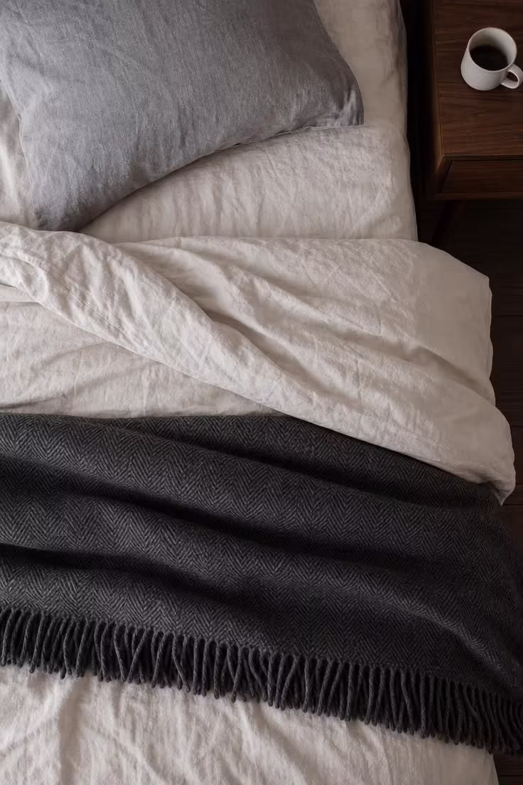 13 Cozy Bedroom Ideas for Women Over 40 You Will Love 3 An overhead view of a bed with stone-washed ivory cotton sheets and a charcoal lambswool herringbone throw in cool overcast light.
