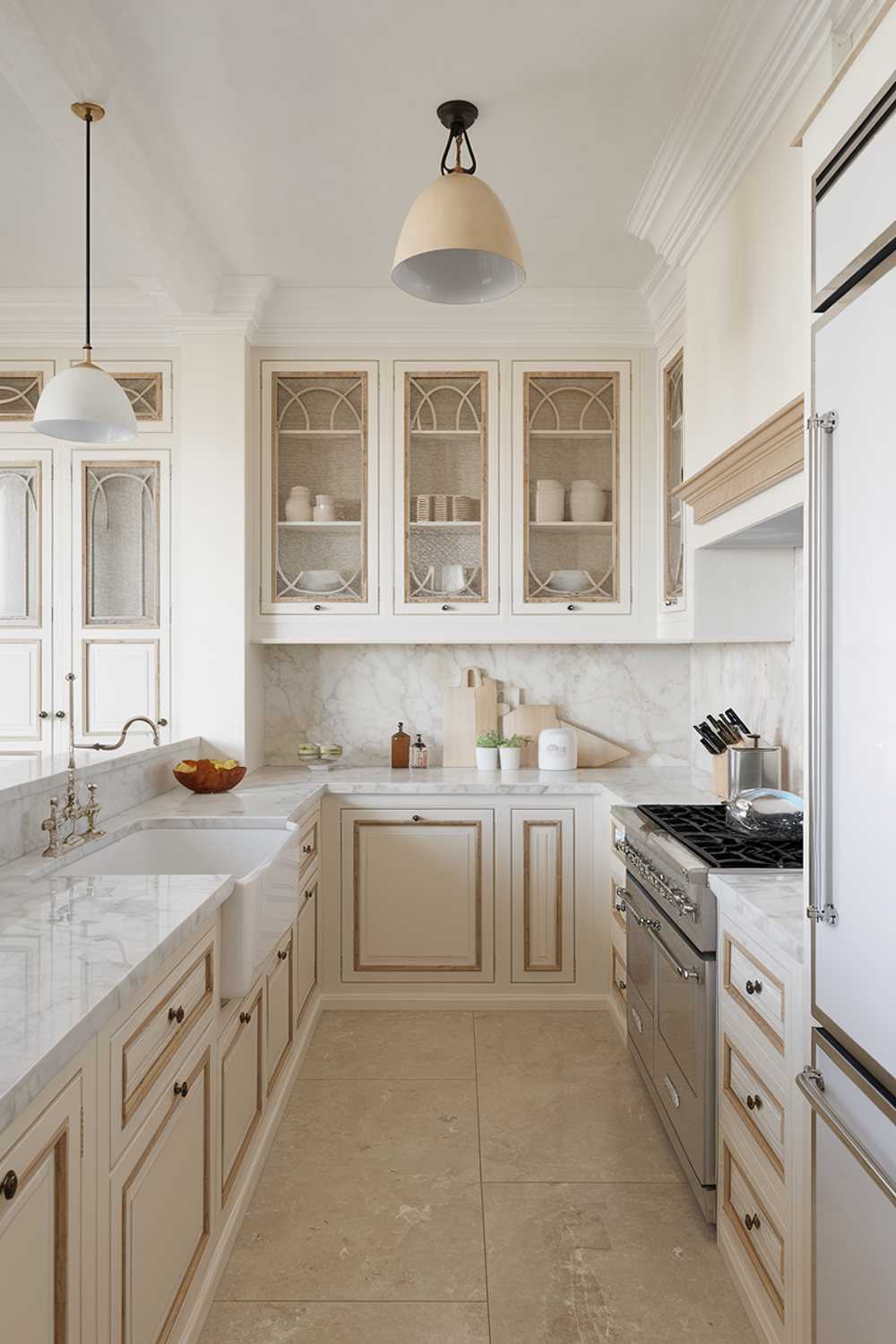 A white and beige kitchen design. The kitchen has a marble countertop and features a double sink, a stove, and a refrigerator. There are wooden cabinets with beige doors and white frames. The floor is beige tile. There is a white pendant light above the sink and a beige pendant light above the stove.