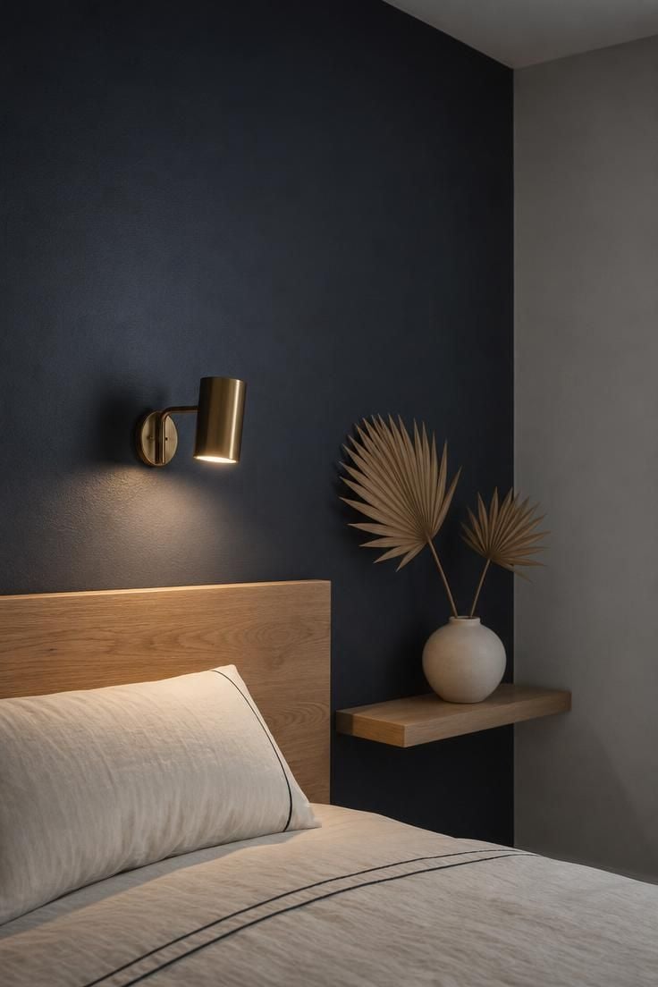 Bedroom accent wall in deep slate blue behind a white oak headboard with a brass sconce casting warm focused light, adjacent walls in pale cool gray for depth contrast.