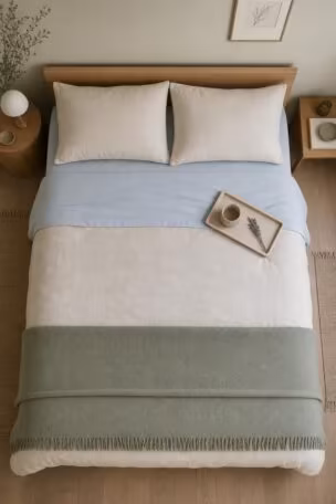 An overhead view of a neatly made bed with pale blue and sage bedding on neutral linen, with a small oak tray holding a ceramic cup and dried lavender.