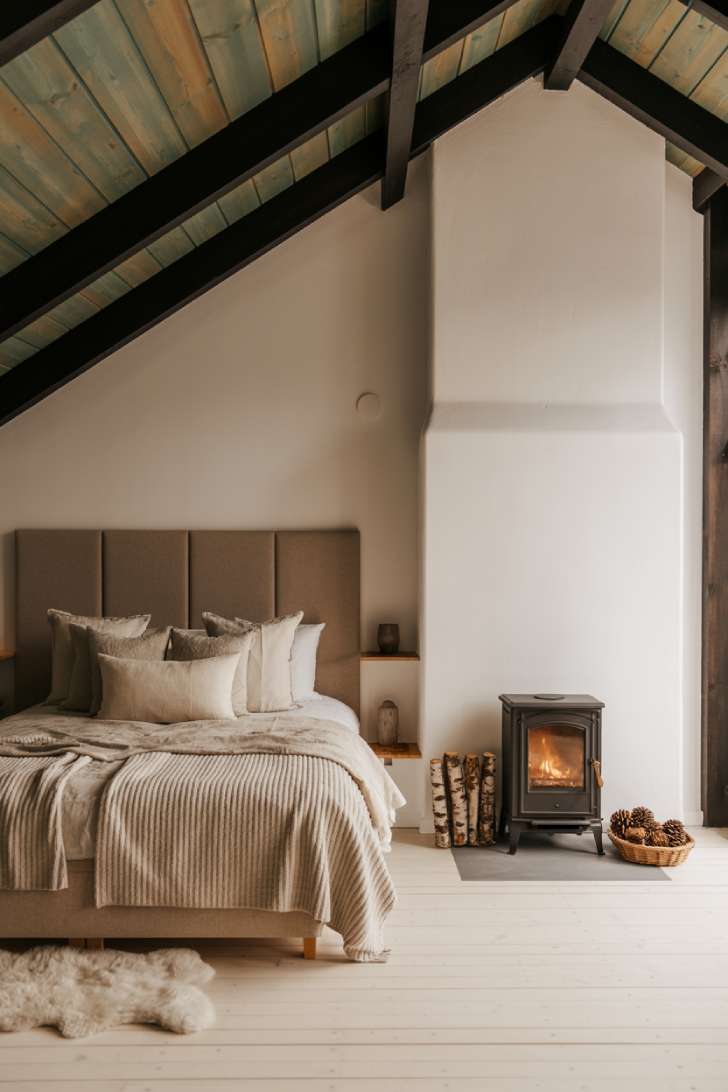 A cozy hygge-inspired attic bedroom with sloped dark pine ceiling, alcove bed, cream ribbed knit blanket, and a small black wood-burning stove