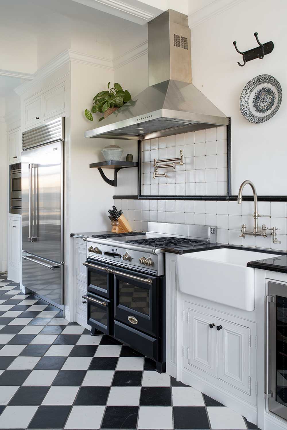 A black and white kitchen with a stylish and highly detailed design. The kitchen has a gas range with four burners, a vent hood above it, and a double oven below. There's a farmhouse sink with a stainless steel faucet next to it. On the left, there's a refrigerator with a stainless steel door. Above the refrigerator, there's a shelf with a potted plant. The floor is made of black and white checkered tiles. The walls are adorned with a few hooks and a decorative plate. The lighting is bright and clean.