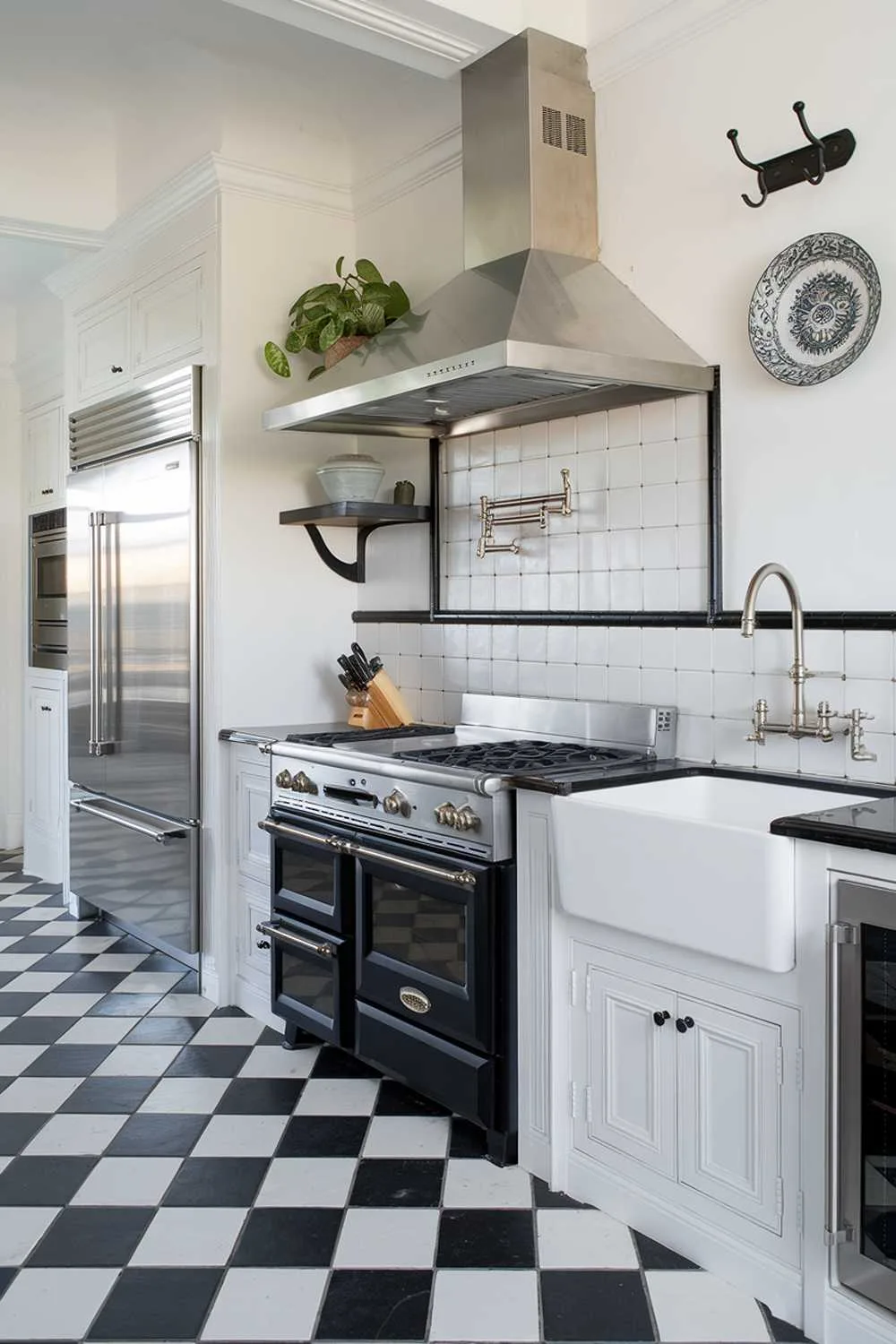 23 Stunning Black and White Kitchen Ideas for Timeless Style 4 A black and white kitchen with a stylish and highly detailed design. The kitchen has a gas range with four burners, a vent hood above it, and a double oven below. There's a farmhouse sink with a stainless steel faucet next to it. On the left, there's a refrigerator with a stainless steel door. Above the refrigerator, there's a shelf with a potted plant. The floor is made of black and white checkered tiles. The walls are adorned with a few hooks and a decorative plate. The lighting is bright and clean.