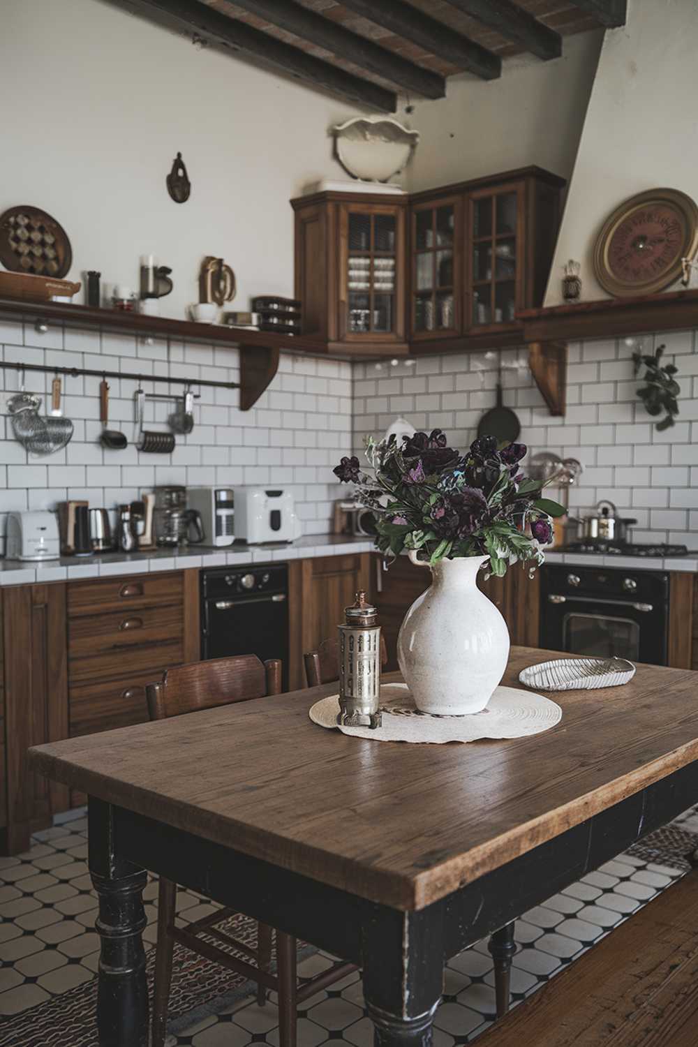 A black and white kitchen decor. The kitchen has a rustic charm with its wooden cabinets, subway tiles, and vintage appliances. There's a large wooden table in the middle of the room. On the table, there's a white vase with dark purple flowers and a few kitchen utensils. The walls have a few hanging items.