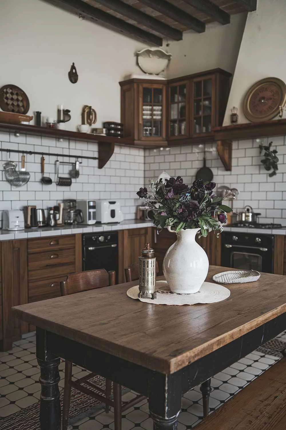 23 Stunning Black and White Kitchen Ideas for Timeless Style 5 A black and white kitchen decor. The kitchen has a rustic charm with its wooden cabinets, subway tiles, and vintage appliances. There's a large wooden table in the middle of the room. On the table, there's a white vase with dark purple flowers and a few kitchen utensils. The walls have a few hanging items.