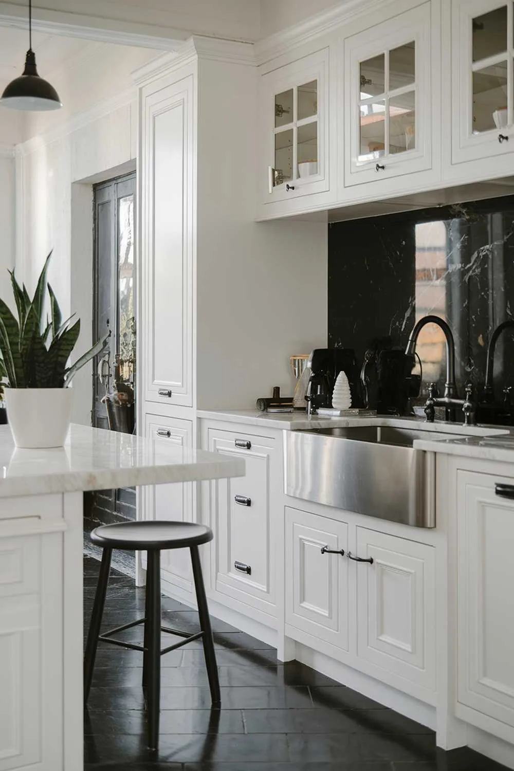 23 Stunning Black and White Kitchen Ideas for Timeless Style 10 A white kitchen with black accents. The kitchen has a marble countertop, a stainless steel sink, and a black faucet. There is a white cabinet with black handles and a black backsplash. There is a black barstool and a white potted plant. The floor is made of black and white tiles. The lighting is bright.