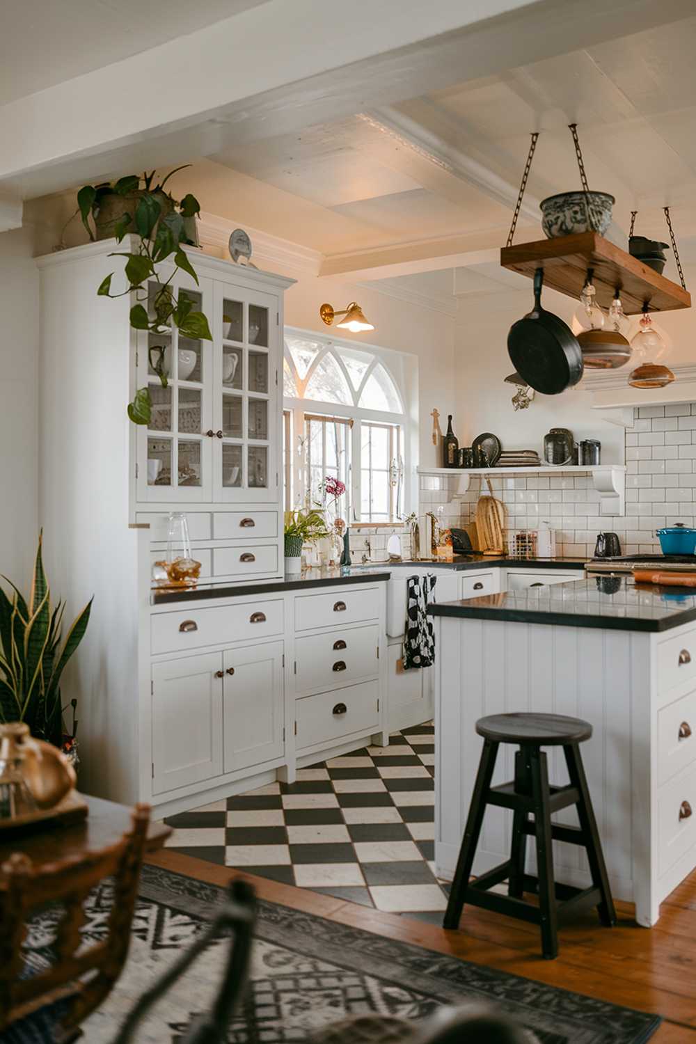 A white kitchen with black accents. The kitchen has a white cabinets, a white island with black countertops, and a white backsplash with black tiles. There is a black stool next to the island. A pot and a pan are hanging above the island. A plant is placed on the white cabinets. The floor is made of black and white checkered tiles. The lighting is warm and inviting