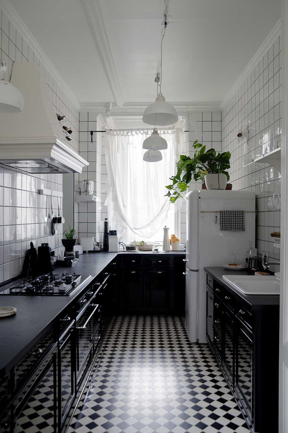  stylish black kitchen with white accents. The kitchen has a black countertop, black cabinets, and a black stove. There is a white fridge and a white sink. There are white lighting fixtures and a white shelf. The floor is black and white checkered. The room has a window with a curtain. There is a potted plant near the fridge.