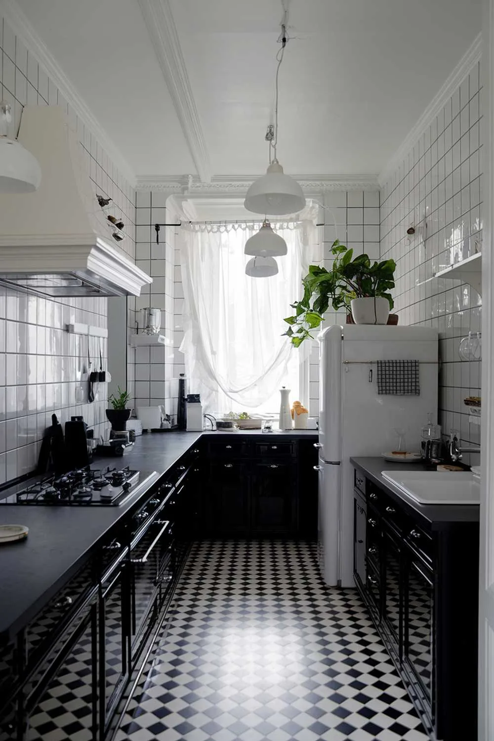 23 Stunning Black and White Kitchen Ideas for Timeless Style 22 stylish black kitchen with white accents. The kitchen has a black countertop, black cabinets, and a black stove. There is a white fridge and a white sink. There are white lighting fixtures and a white shelf. The floor is black and white checkered. The room has a window with a curtain. There is a potted plant near the fridge.