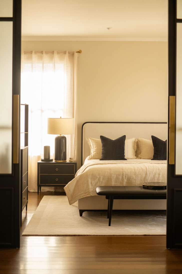 A bedroom featuring a black leather bench, a black floor lamp with a cream shade, and brass nightstand hardware