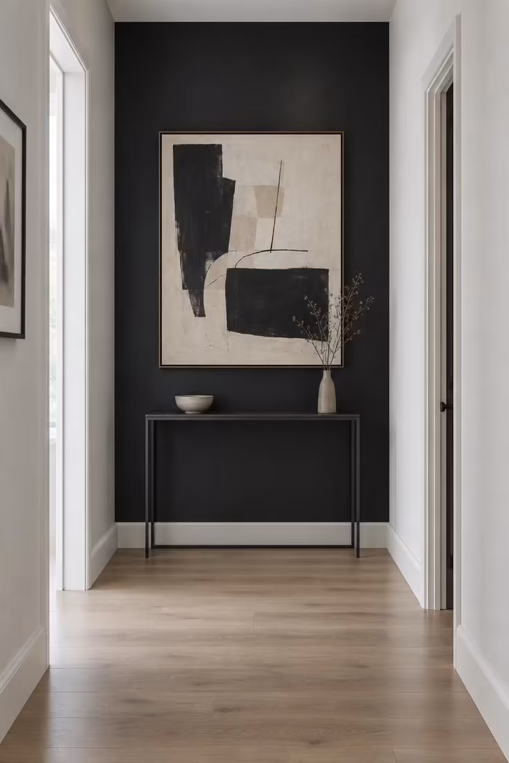 A hallway viewed straight on toward a single black accent wall at the end, with one large framed artwork and a slim console beneath it in cool overcast light.