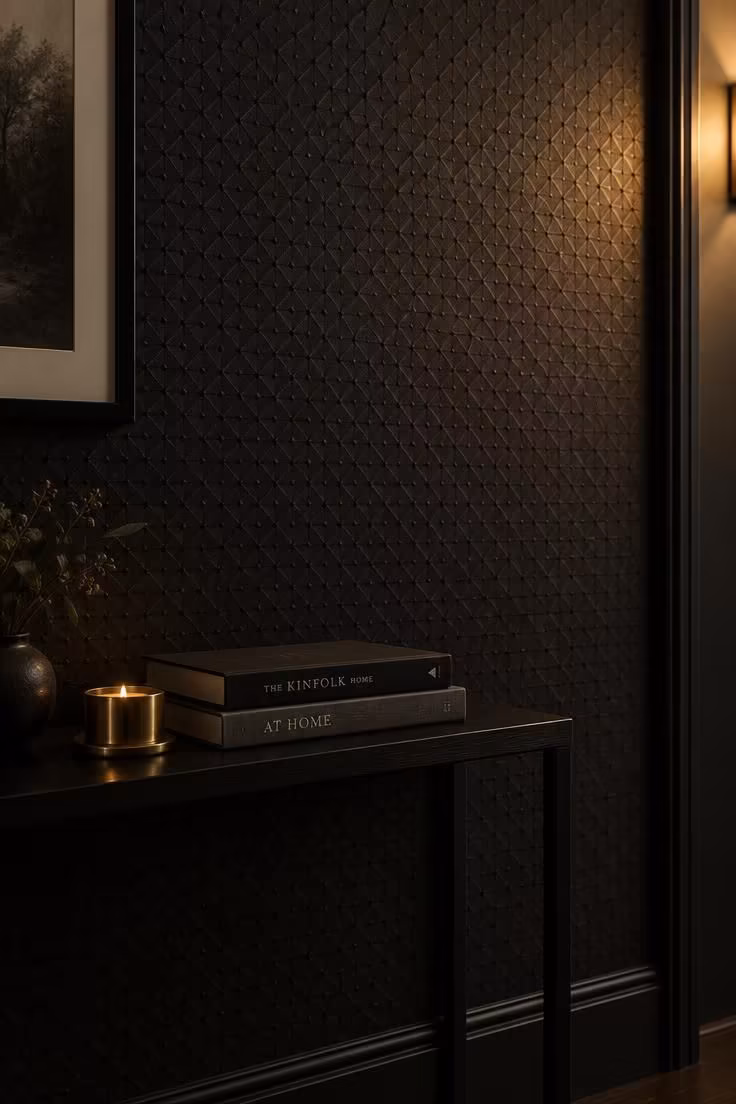 A close view of black-on-black patterned wallpaper with a small geometric repeat, lit by warm sconce light that reveals the texture of the wall.