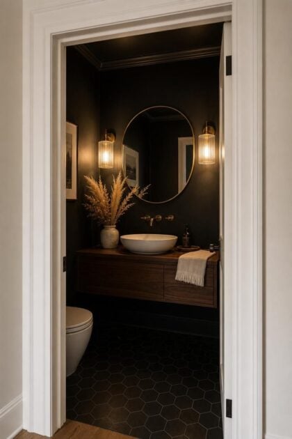 11 Bold Black Powder Room Ideas for a Moody Retreat Cozy black powder room seen through white doorway frame with brass sconces, floating walnut vanity, and warm golden lighting