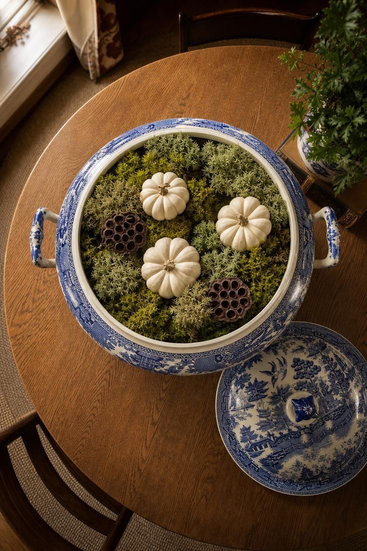 11 Cozy Blue and White Fall Decor Ideas for Warmth 4 A vintage Blue Willow tureen filled with green moss, miniature white pumpkins, and dried lotus pods on an oak dining table viewed from above.
