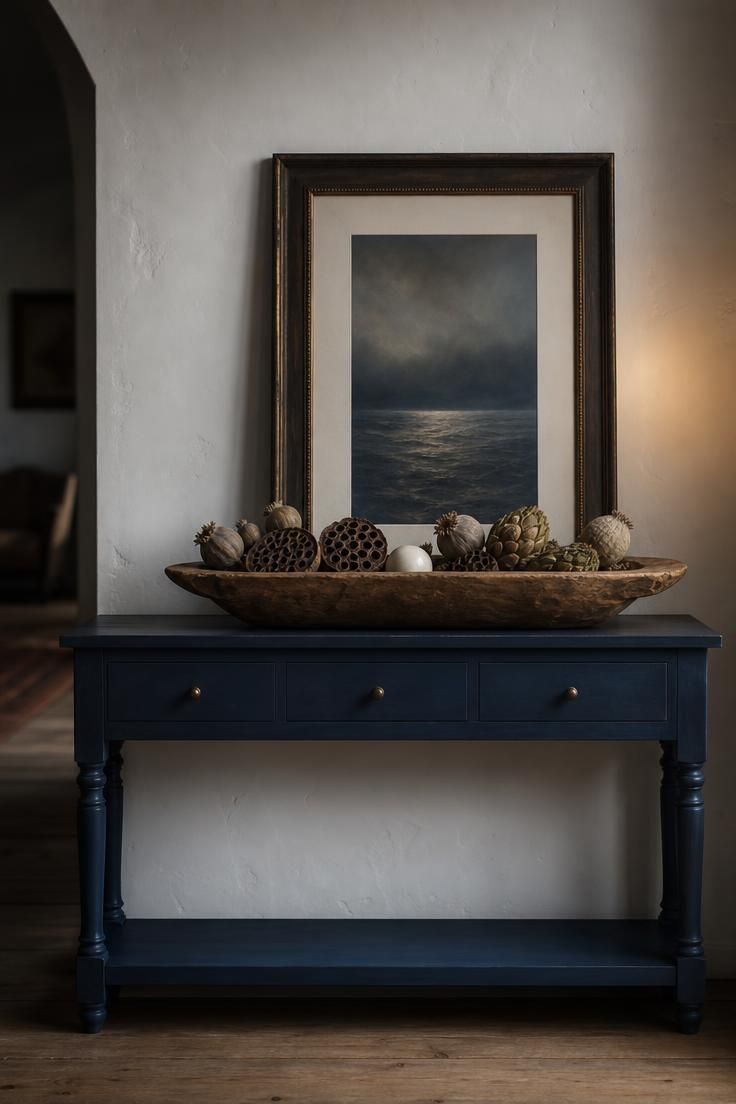 11 Cozy Blue and White Fall Decor Ideas for Warmth 10 A moody indigo console table with a raw wood dough bowl filled with dried seed pods and a white ceramic sphere, a blue seascape print leaning behind.