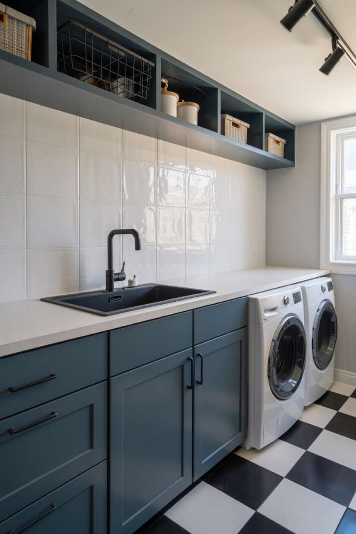Slate blue flat-panel cabinets with matte black hardware, black-framed window, and black and white checkered floor in a modern laundry room