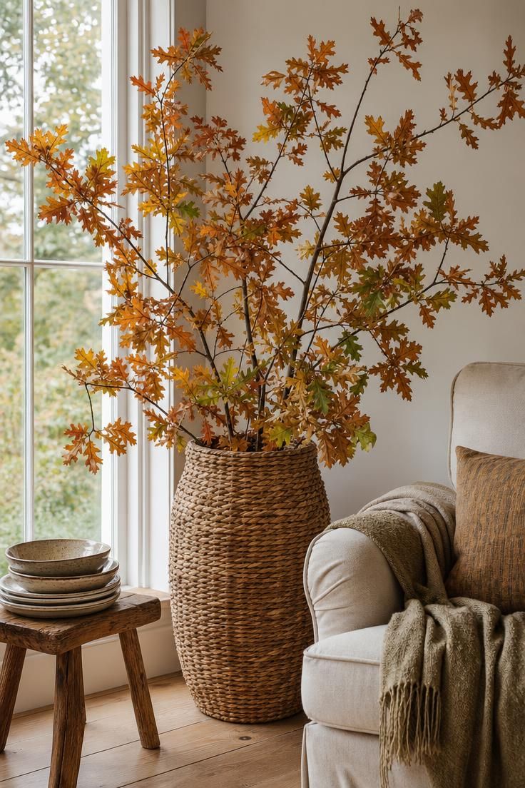 Foraged oak branches with turning leaves in a tall seagrass basket beside a window in soft daylight.