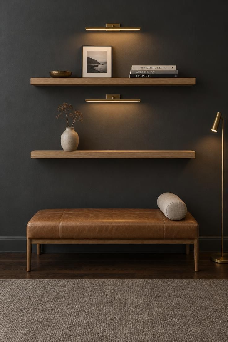 15 Cozy Classy Man Cave Ideas to Make Your Retreat 3 Charcoal gray wall with two floating white oak shelves lit by brass picture lights, holding a bronze dish, framed black-and-white photograph, art books, and a ceramic vase above a low tan leather bench with a bolster pillow.