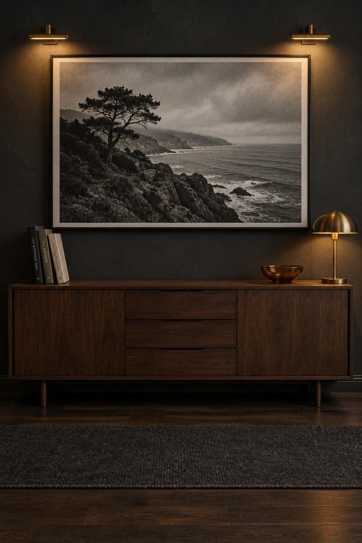 15 Cozy Classy Man Cave Ideas to Make Your Retreat 11 Large black-and-white photograph on a charcoal wall above a low walnut sideboard, lit by two brass picture lights, with clothbound books and an amber glass bowl on the sideboard below.