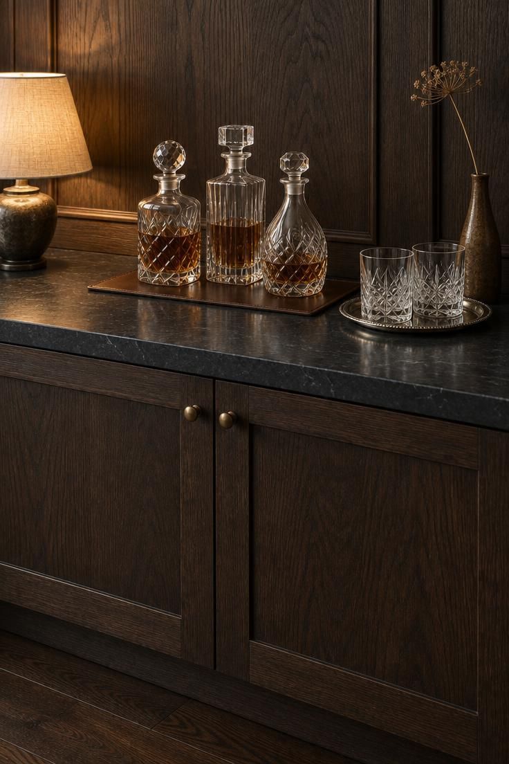 15 Cozy Classy Man Cave Ideas to Make Your Retreat 14 Low cabinet with a honed black stone top against dark wood paneling, holding three crystal decanters on a leather mat, two old-fashioned glasses on a silver tray, and a narrow vase with a dried stem, lit by a small lamp.