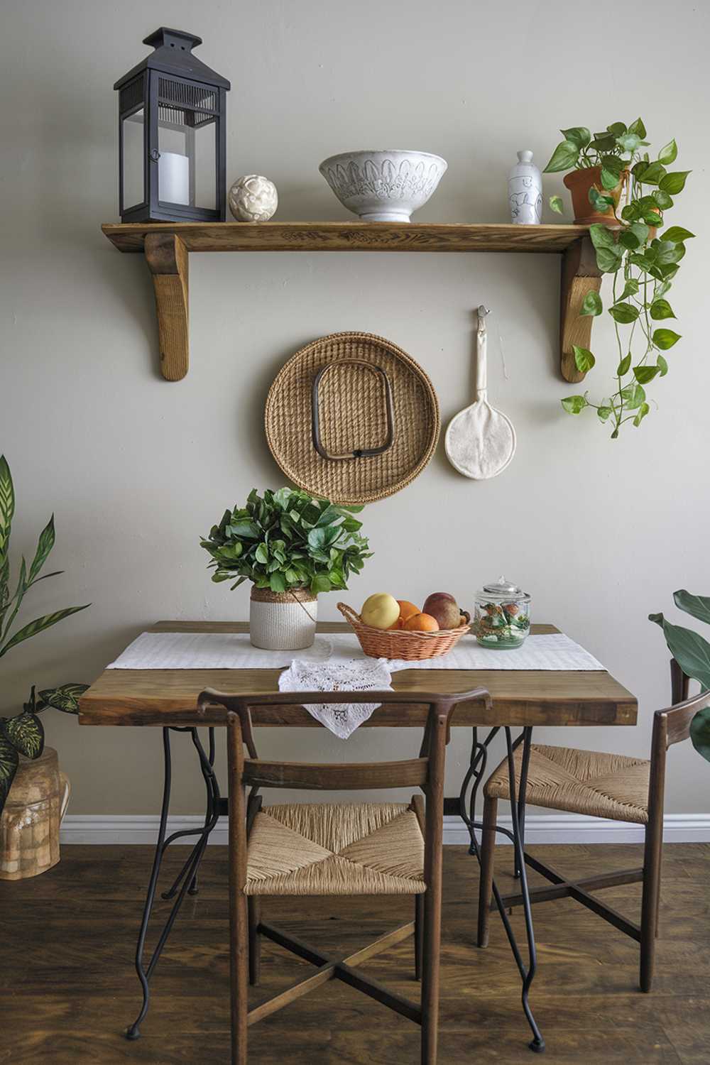 A coastal farmhouse kitchen decor. There's a wooden table with a white tablecloth and a few chairs. On the table, there's a green plant, a basket of fruits, and a few decorative items. The wall behind the table has a wooden shelf with a few decorative items, including a lantern, a white bowl, and a few green plants. The floor is made of wood.