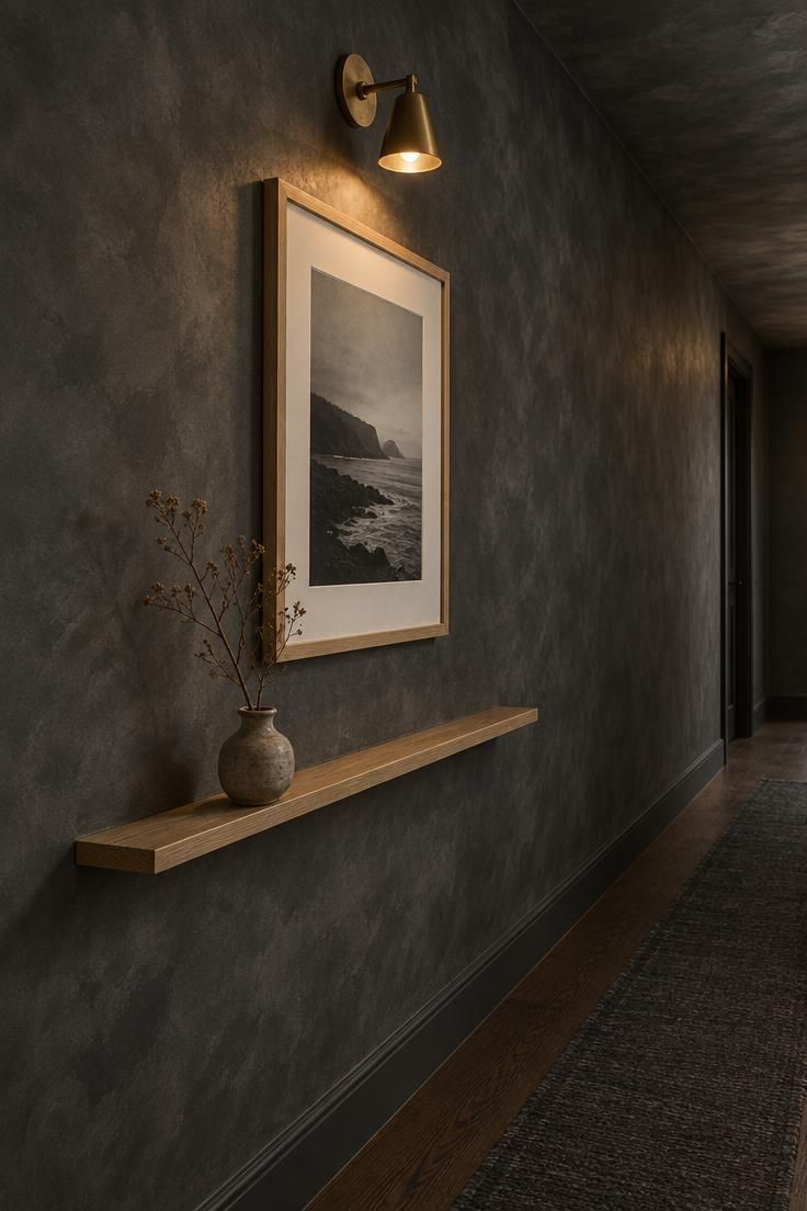 A charcoal limewash hallway wall with one large pale oak frame, a black-and-white print, and a single brass sconce above it.