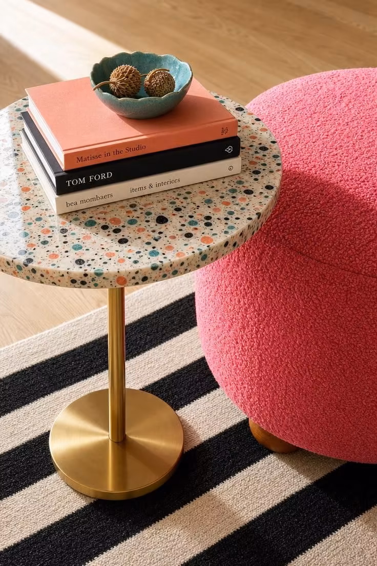 Round terrazzo side table with brass stem next to hot pink ottoman on striped rug with art books.