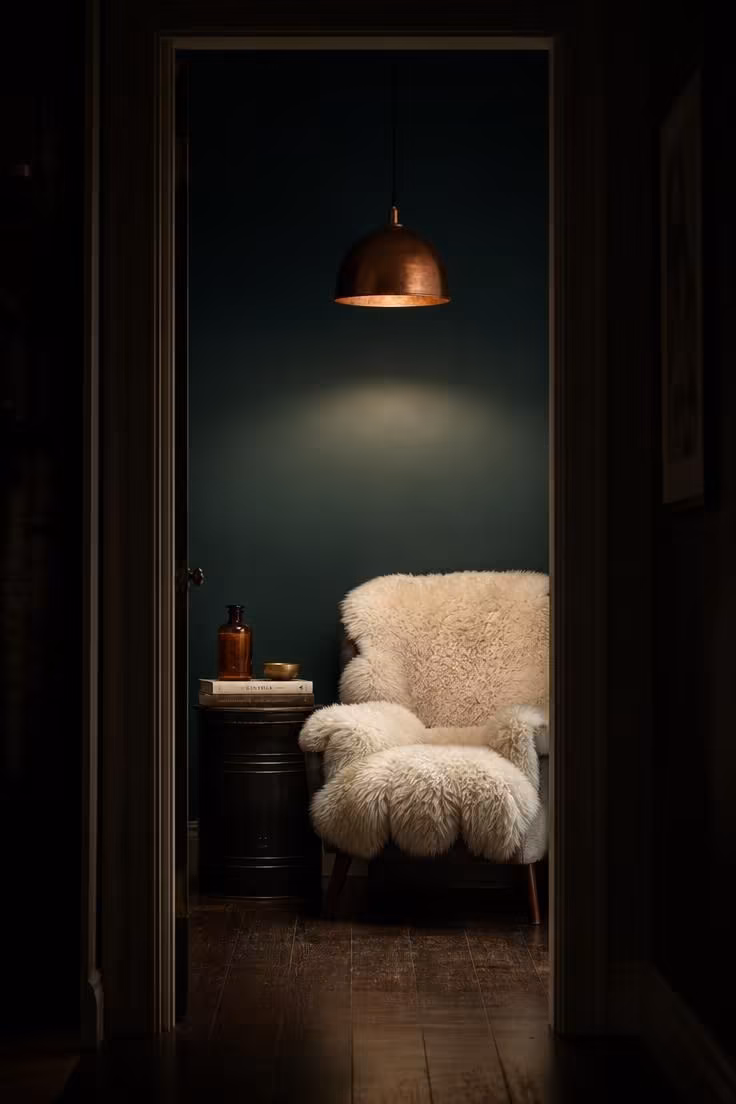 Deep teal wall with copper dome pendant over sheepskin chair and black metal side table with amber glass bottle.