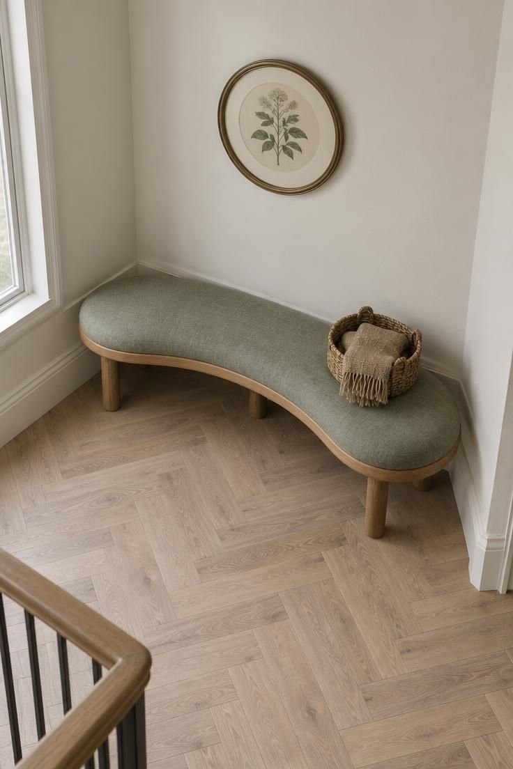 An overhead view of a curved oak bench upholstered in sage green linen tucked into a widening hallway corner, with a woven basket and jute throw on the seat and a round botanical print on the wall above.