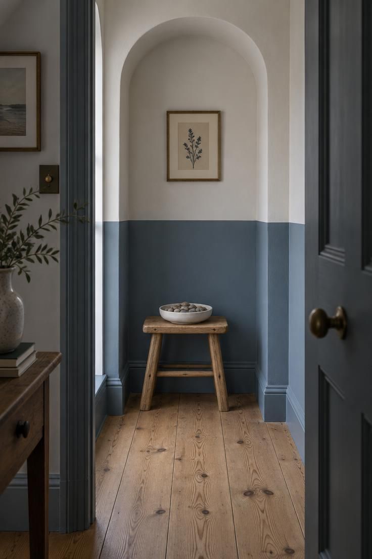 A small hallway corner niche with a dusty blue lower half and warm white upper half, holding a low raw oak stool with a round white ceramic dish of river stones above wide pine flooring.