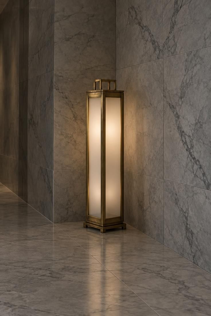 A tall brass floor lantern with frosted glass panels standing diagonally in a hallway corner where two pale gray marble-tiled walls meet above large-format marble flooring.