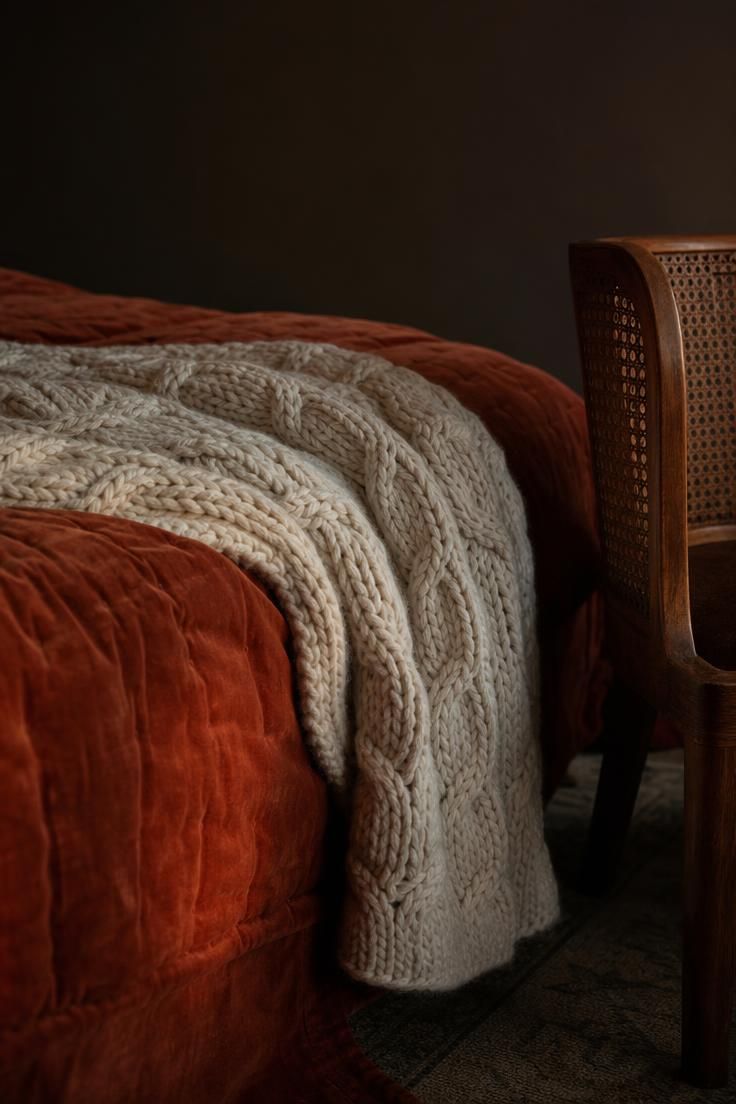 Chunky cream knit throw draped over a terracotta velvet quilt with the edge of a cane-side chair entering the frame.