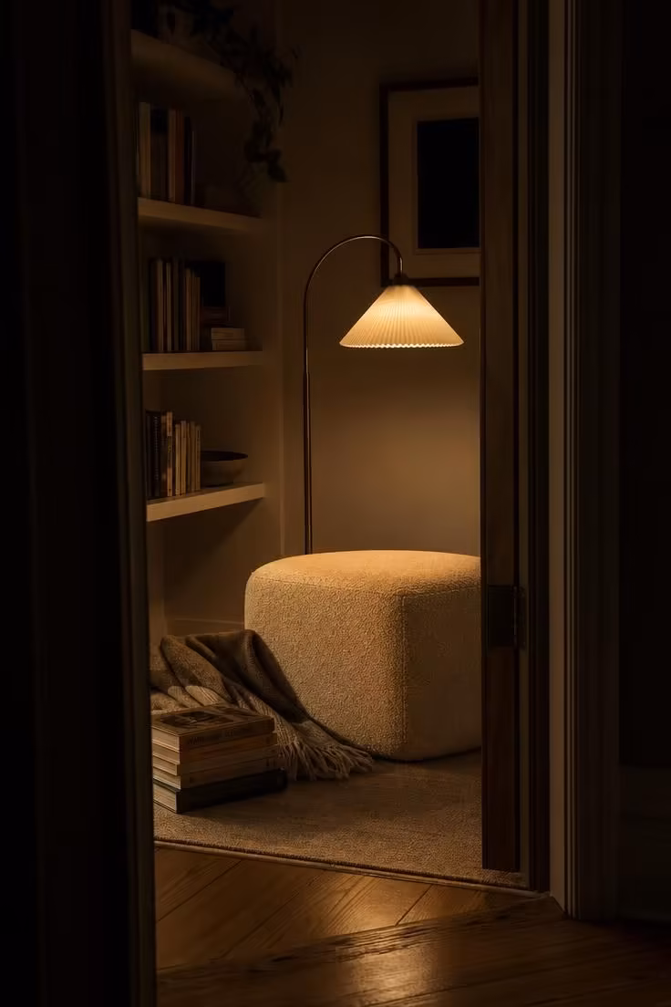 11 Cozy Eclectic Living Room Ideas Full of Warmth and Charm 5 Cream bouclé ottoman beside an arched brass floor lamp in a moody, low-lit reading nook.