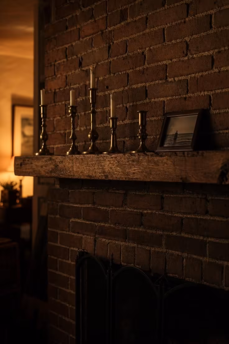 11 Cozy Eclectic Living Room Ideas Full of Warmth and Charm 10 Brick fireplace mantel styled with mismatched brass candlesticks in warm, moody evening light.