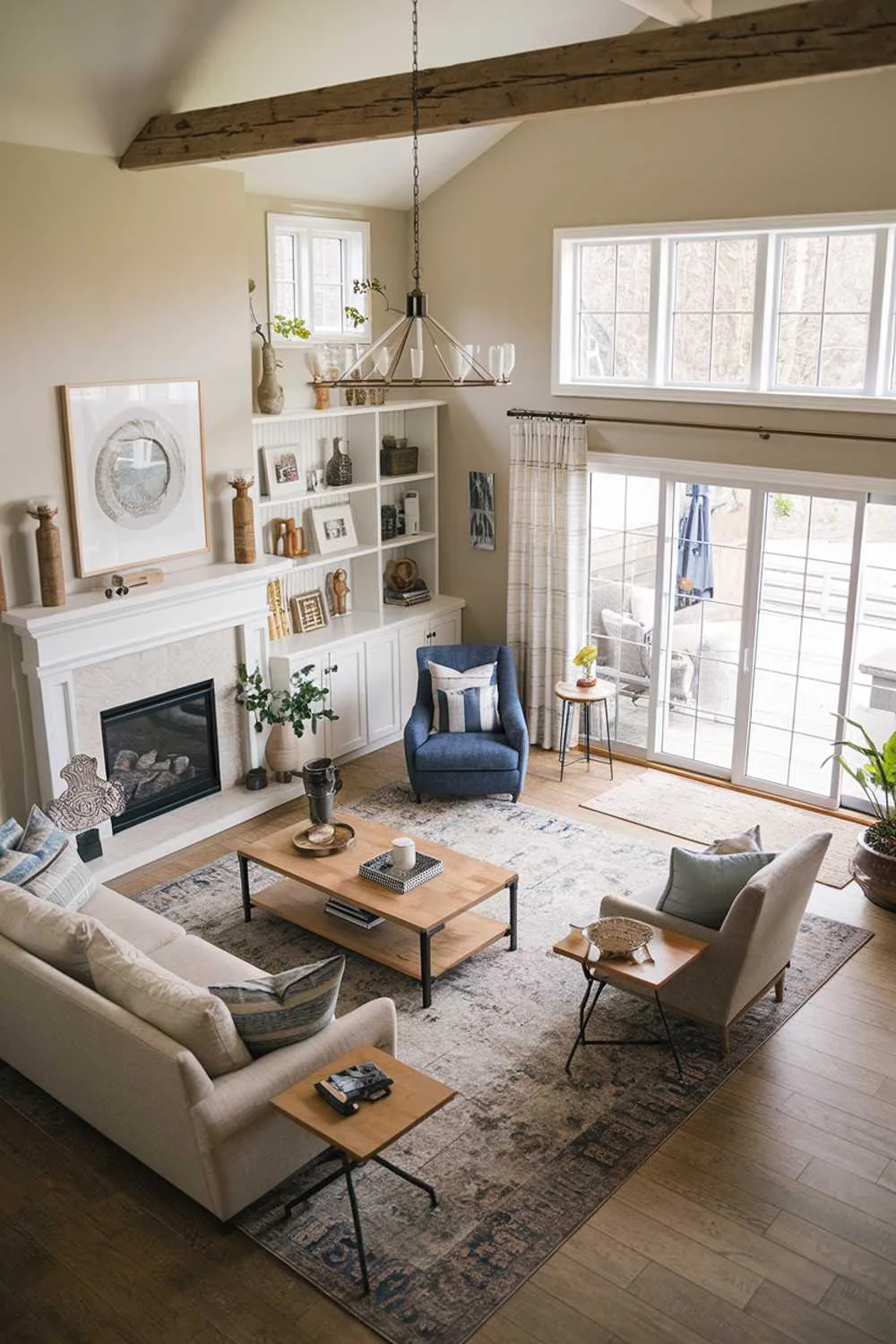 21 Cozy Farmhouse Living Room Ideas to Warm Your Home 4 A cozy farmhouse living room design and decor. The room has a rustic charm with modern elements. There's a beige sofa, a blue armchair, a wooden coffee table, and a white bookshelf. The walls are adorned with decorative items. A large area rug covers the wooden floor. A sliding glass door leads to an outdoor space.