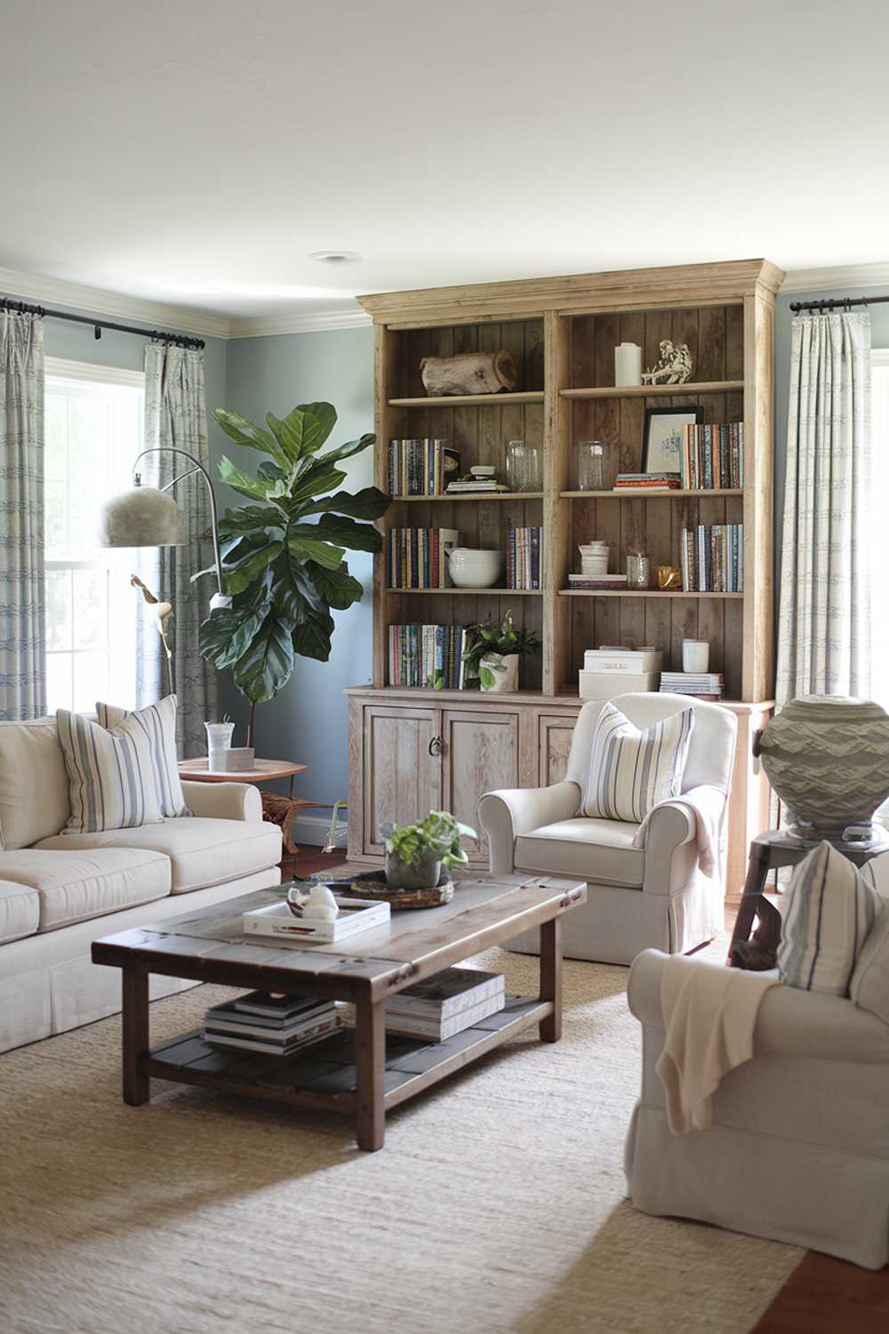  A cozy farmhouse living room with a rustic-chic aesthetic. The room has a beige sofa with patterned pillows, a dark wooden coffee table, and a beige armchair. There's a wooden bookshelf filled with books and decor. A large potted plant sits on the floor. The room has a beige area rug. The walls are painted in a light blue hue.