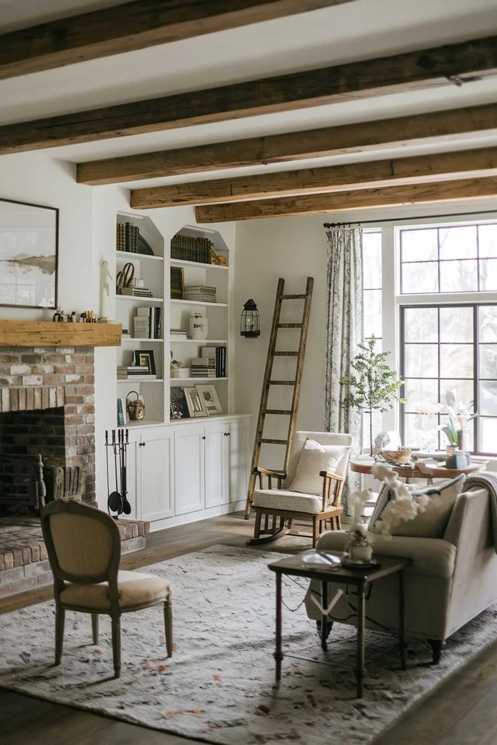 21 Cozy Farmhouse Living Room Ideas to Warm Your Home 21 A cozy farmhouse living room with a rustic charm. The room has a wooden beam ceiling, a brick fireplace with a mantel, and a plush rug in front of the fireplace. There is a vintage wooden chair near the fireplace. A sofa, a rocking chair, and a side table are placed near the window. A ladder leans against the wall near the window. The room has a few decorative items, including a lantern and a small tree. The walls are adorned with shelves holding books and decor items.