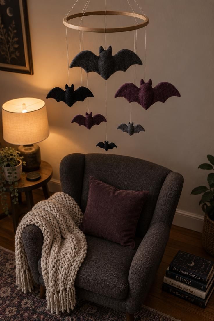 10 Cute Halloween Decor Ideas for a Warm, Friendly Home 6 A felt bat mobile with graduated charcoal and plum bats hanging from a wooden hoop above a reading chair, lit by a warm lamp against a pale wall.