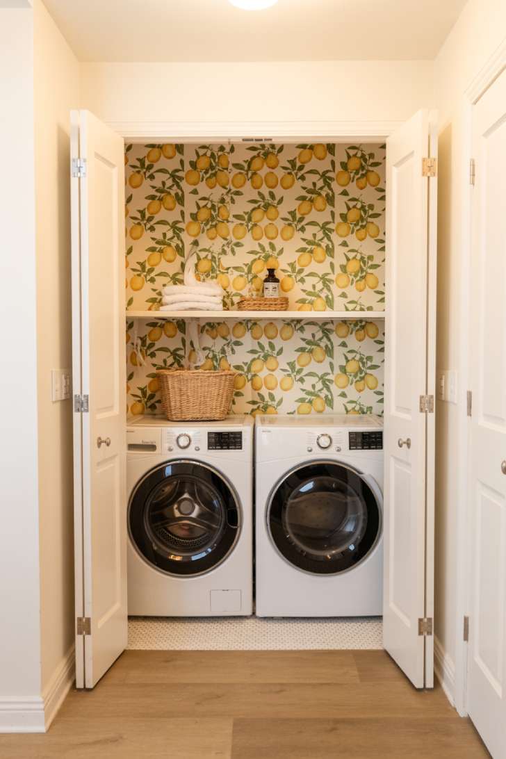 A small laundry closet with lemon-print wallpaper, a stacked washer and dryer, and a floating shelf.