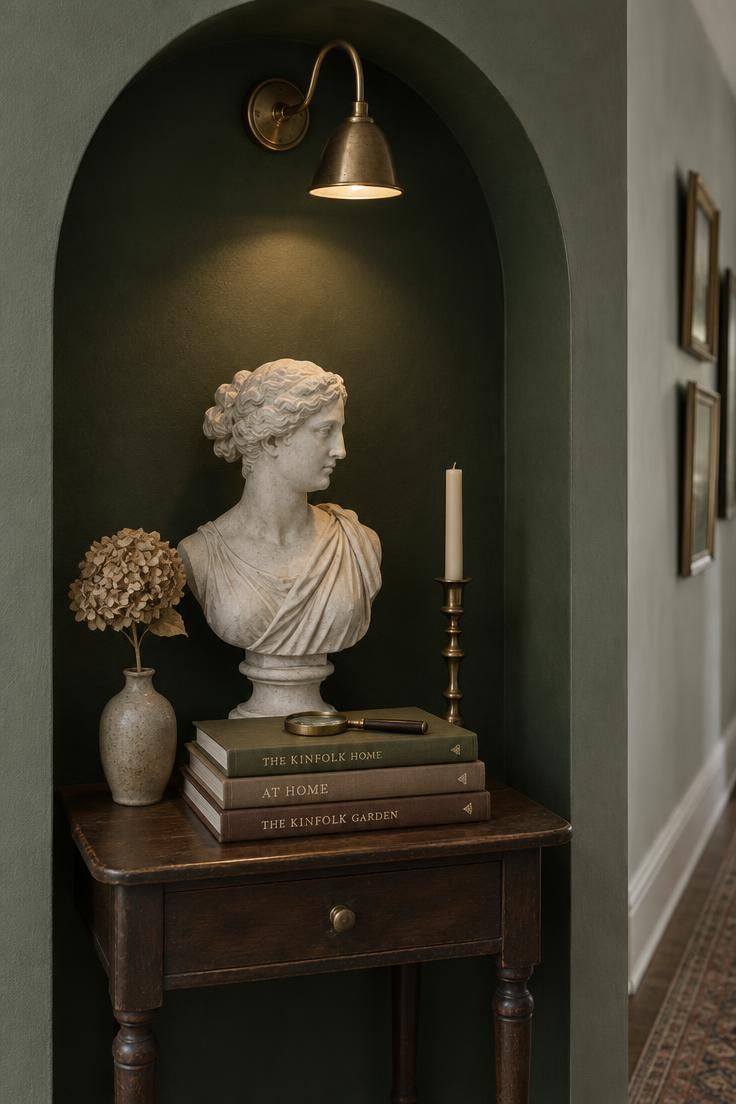 A deep arched alcove in a forest green hallway wall holding a plaster bust, cloth-bound books, and a brass candlestick, lit by a brass gooseneck lamp above the arch.