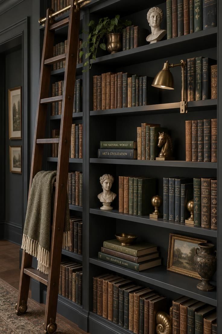A floor-to-ceiling charcoal bookshelf built into a hallway recess, packed with vintage books and small plaster busts, with a slim rolling ladder on a brass rail.