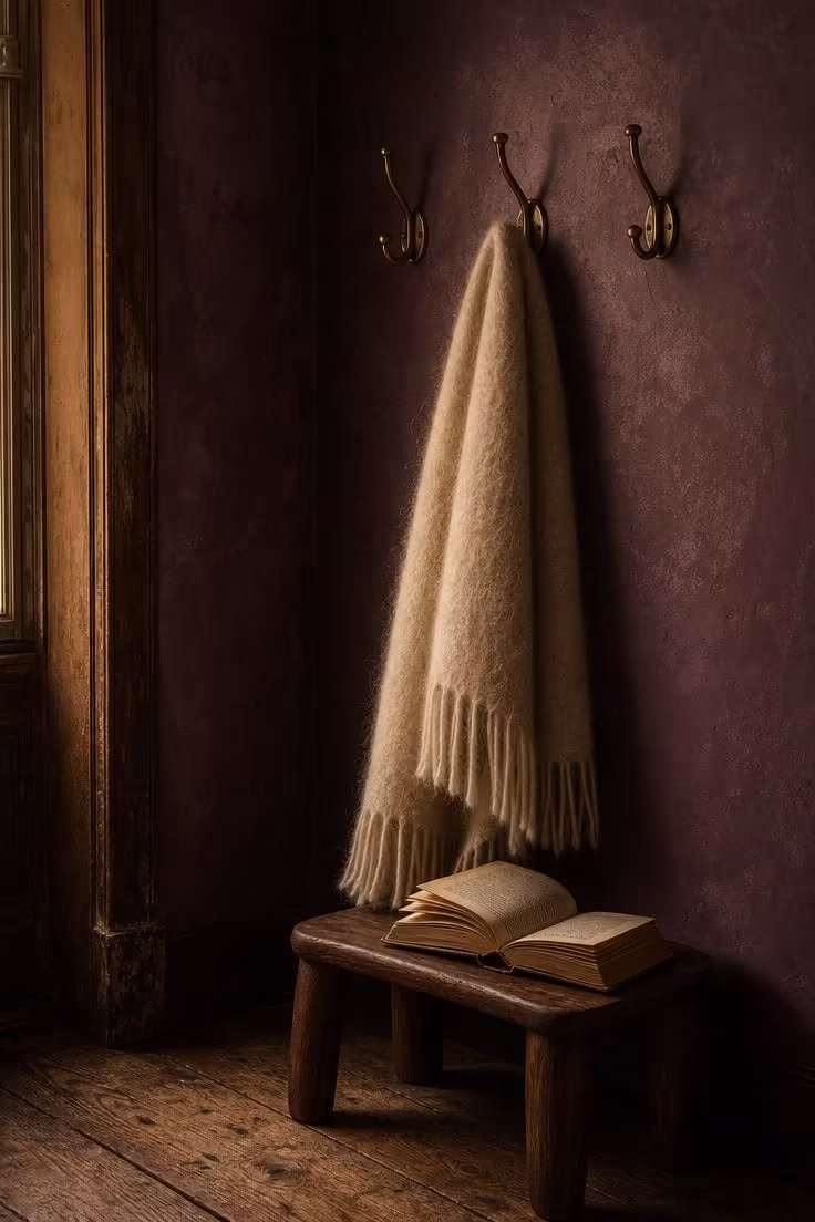13 Rich Dark Academia Reading Nook Ideas Worth Copying 4 Dark plum limewash wall with brass coat hooks holding a cream mohair throw, a walnut stool, and an open book.