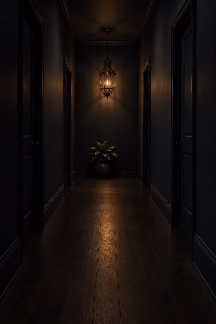 14 Warm Dark Hallway Ideas to Make a Bold Statement 13 Long corridor with walls, doors, and skirting in one deep tone, a single blackened metal lantern glowing at the far end, and a low plant for scale.