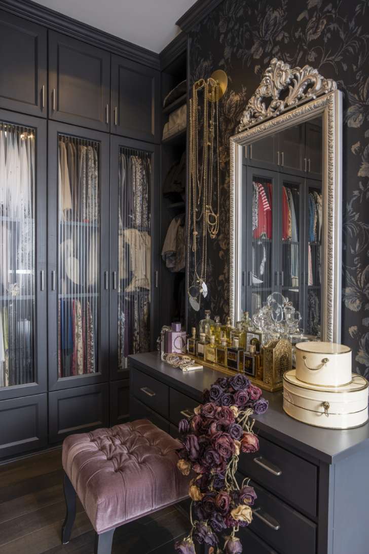 Dark maximalist dressing room with fluted glass wardrobes, baroque mirror, dark floral wallpaper, and vintage perfume display