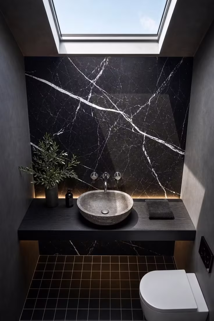 Dark marble slab accent wall with backlit floating shelf and stone vessel sink under skylight light