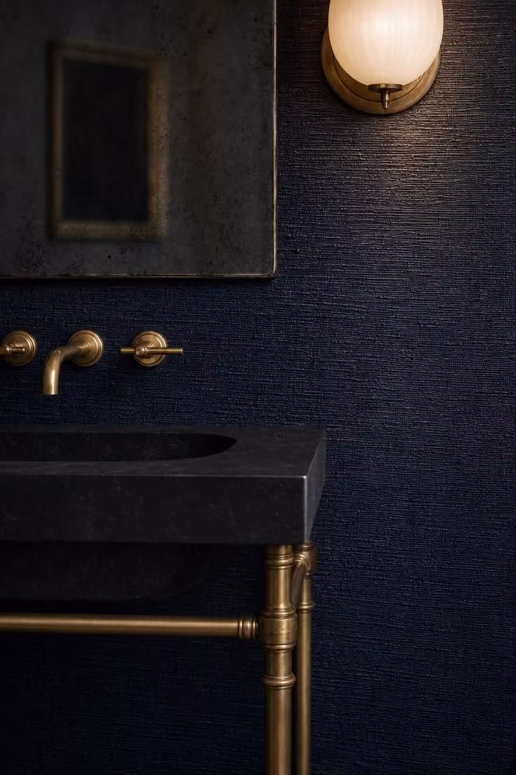 Inky navy grasscloth wall with stone console sink on aged brass legs and antiqued mirror